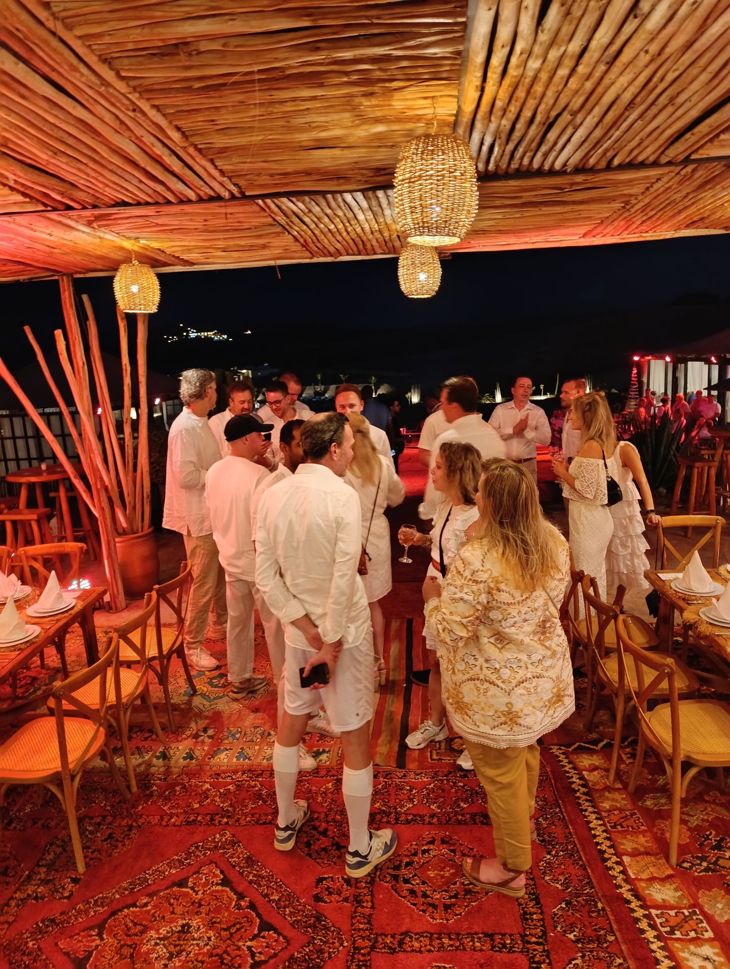 Group of people in white attire socializing under a bamboo roof with warm lighting and decorative rugs.